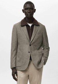 Sako s vzorem psí zub v hnědé a béžové barvě, s hnědým límcem, dvěma předními kapsami a zapínáním na knoflíky. Nahoře je nošeno přes svetr na zip.