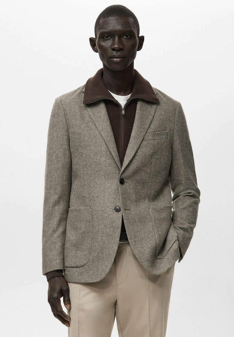 Sako s vzorem psí zub v hnědé a béžové barvě, s hnědým límcem, dvěma předními kapsami a zapínáním na knoflíky. Nahoře je nošeno přes svetr na zip.