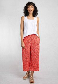 White sleeveless top with lace detailing, paired with red floral wide-leg pants. Model wears tan espadrille sandals. Textured background.