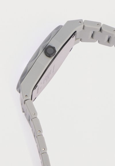 Πλευρική όψη ενός γκρι μεταλλικού ρολογιού χειρός με υφασμένο στεφάνι και μαύρο κουμπί κορώνας στο κουτί.