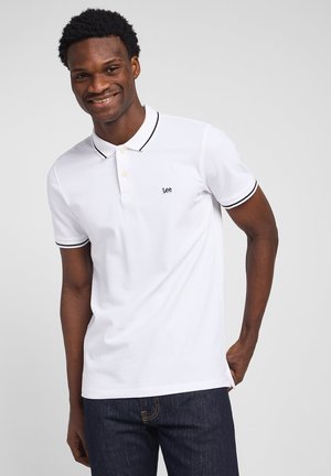 Lächelnder Mann, der ein weißes Poloshirt mit schwarzem Rand und "Lee"-Logo trägt, kombiniert mit dunkelblauen Jeans, vor einem schlichten Hintergrund stehend.