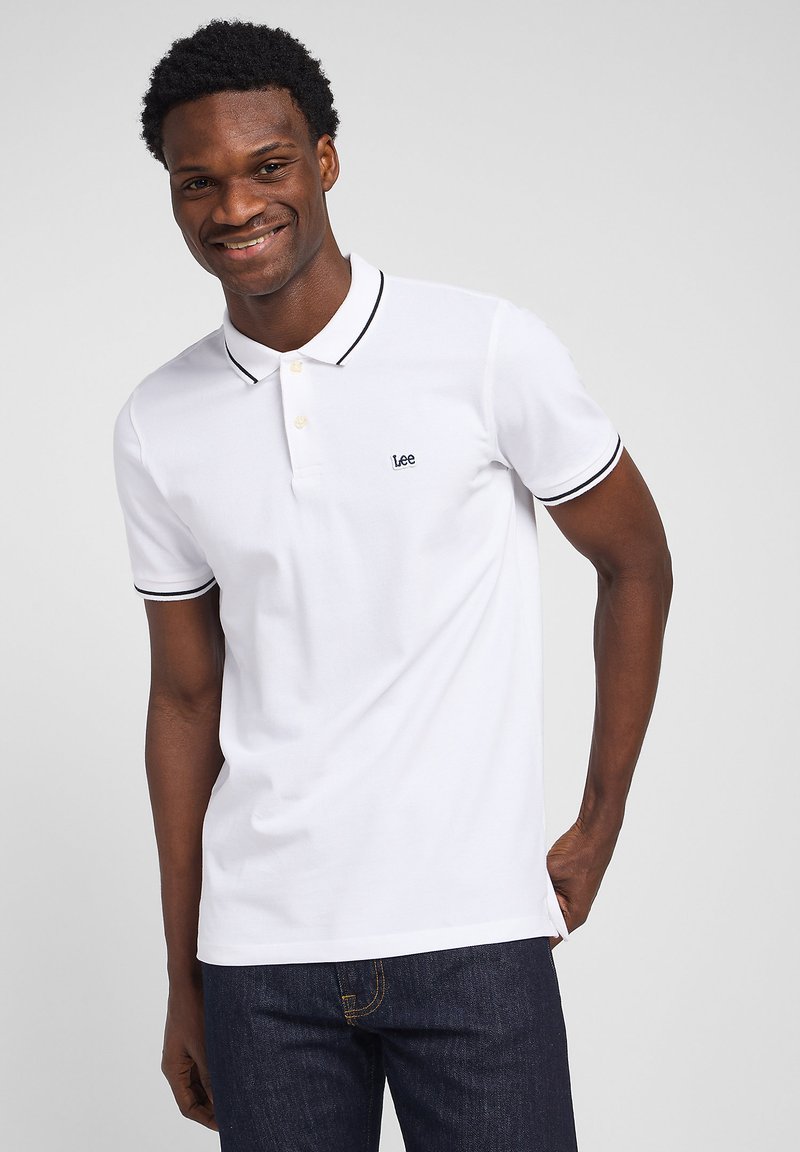 Uśmiechnięty mężczyzna ubrany w biały polo z czarnym obszyciem i logo "Lee", w ciemnoniebieskich dżinsach, stojący na jednolitym tle.