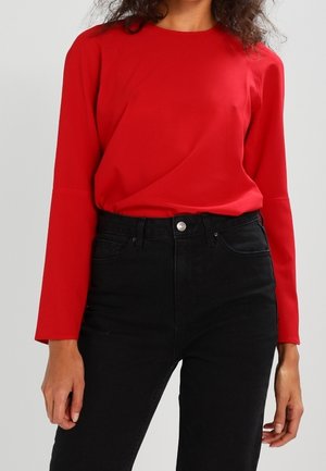 Kobieta ubrana w czerwoną bluzkę z długim rękawem, włożoną do wysokich czarnych dżinsów, na jednolitym tle.