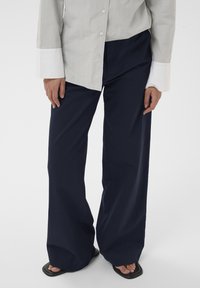 Personne portant un pantalon large bleu marine, une chemise beige à boutons avec des poignets blancs, et des sandales noires à bout carré sur un fond blanc.