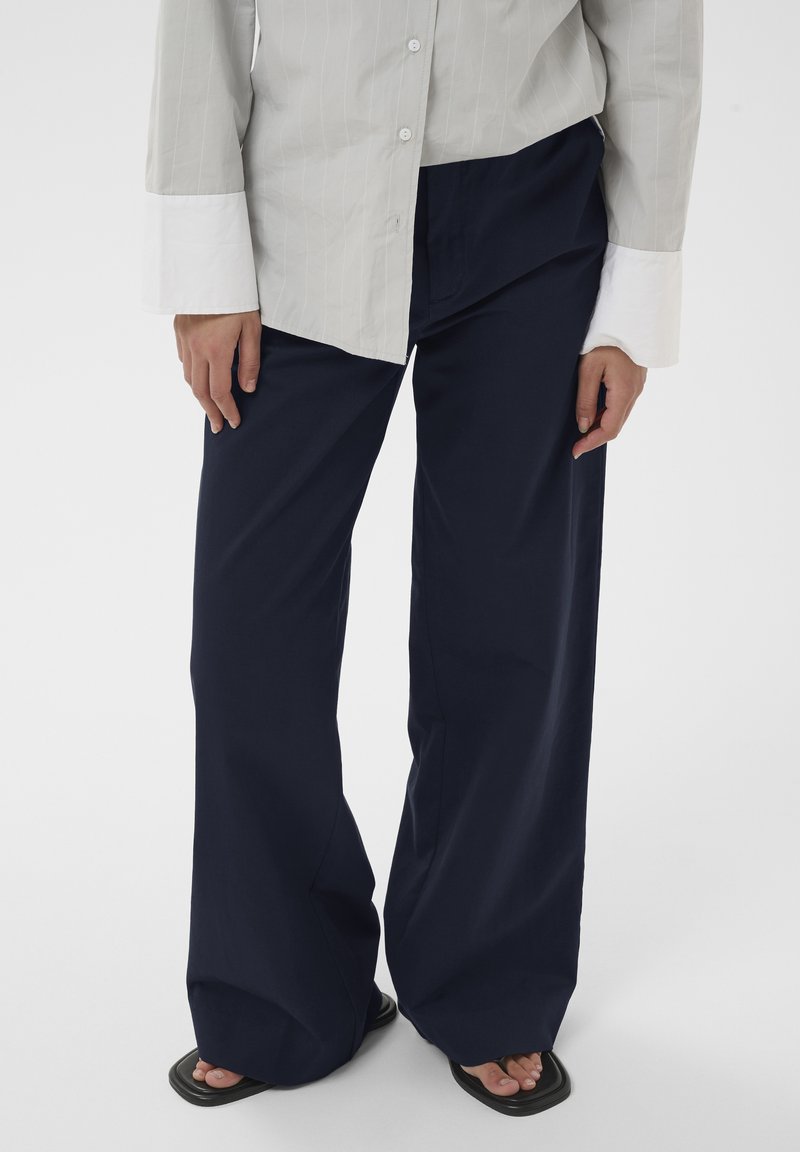 Personne portant un pantalon large bleu marine, une chemise beige à boutons avec des poignets blancs, et des sandales noires à bout carré sur un fond blanc.