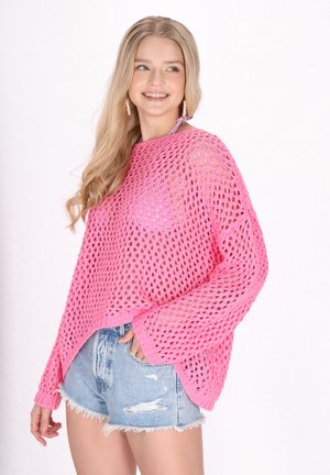 Rosa gehaakte pullover met een groot open breisel en brede mouwen, gedragen over lichtblauwe versleten denim shorts.