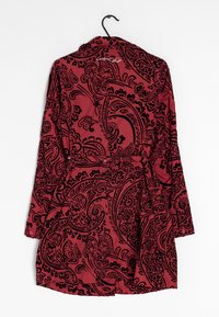 Rode lange mouwenjas met zwarte intrikate paisley-patronen, hangend aan een zwarte hanger tegen een witte achtergrond.