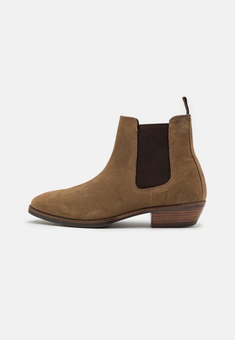 Botte Chelsea en daim marron avec panneau latéral élastique foncé, bout arrondi et talon bas en bois. Finition texturée avec des détails de couture minimaux.