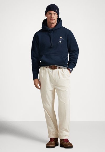 Mann iført marineblå hettejakke med bjørnegrafikk, kremfargede kordfløyelsbukser, brunt belte, brune fjellstøvler, røde sokker, marineblå lue og armbåndsur som står oppreist.