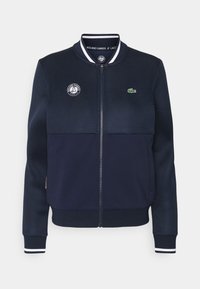 Marinblå bomberjacka med nätpaneler, sidofickor och ribbade mudd. Broderad logotyp på bröstet och randiga kragar. Hela dragkedjan.