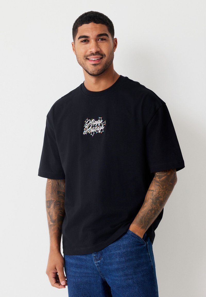 Camiseta de algodón negra con mangas cortas y cuello redondo, con un diseño de texto bordado multicolor en el centro. Se lleva con vaqueros azules.