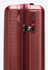 Červený pevný kufr s drážkovaným designem, dvojitými zipy a kombinovaným zámkem. Obsahuje štítek, který uvádí, že materiál je 100% PC.