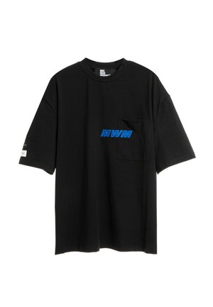 Joven de pie con una camiseta negra de gran tamaño con logo azul, pantalones cortos cargo negros con cremalleras, calcetines negros y zapatos negros con textura.