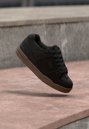 Baskets noires avec semelle et lacets marron, flottant au-dessus d'un trottoir rose avec des marches en béton en arrière-plan.