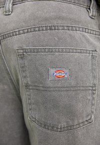Szare dżinsy mają tylnią kieszeń z prostokątną naszywką logo Dickies w kolorze czerwonym, żółtym i niebieskim, podkreślającą funkcjonalny design.