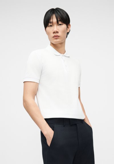 Νέος άντρας με μαύρα μαλλιά, ντυμένος με λευκό πόλο μπλουζάκι με κοντά μανίκια και μαύρο παντελόνι, με τα χέρια στις τσέπες, μπροστά από απλό φόντο.