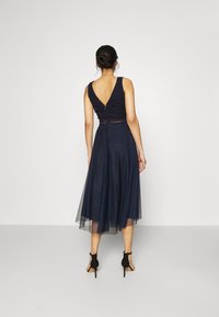 Abito blu navy con corpetto in pizzo e scollatura a V sul retro, vita aderente e gonnellina di tulle fluente fino al ginocchio. Scarpe nere con tacco alto.