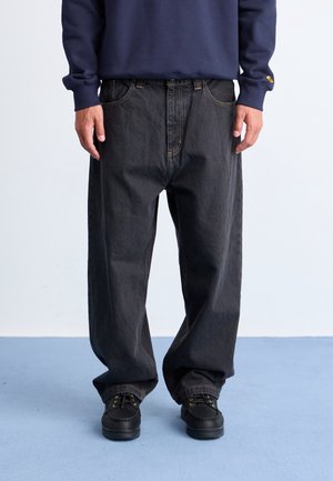 Personne portant un jean noir ample, des chaussures noires et une chemise bleu marine à manches longues, debout sur un sol bleu clair sur fond blanc.