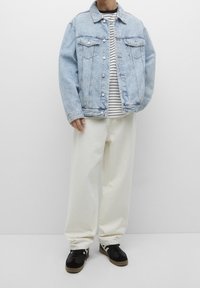 Personne portant une veste en jean bleu clair, un t-shirt à rayures noires et blanches, un pantalon blanc ample et des baskets noires à semelles gomme sur fond blanc.