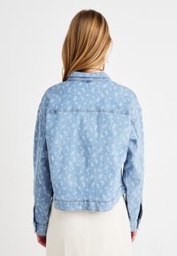 Svetlo modra denim jakna z cvetličnim vzorcem, kratek dizajn, vidno šivanje in gumbi na manšetah. Mehka tekstura z ohlapnim krojem.