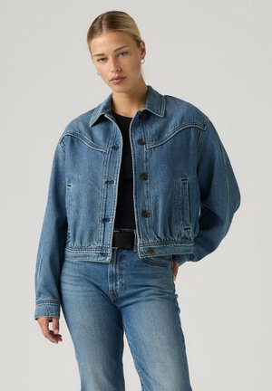 Junge Frau mit blonden Haaren, die eine blaue Jeansjacke und Jeans über einem schwarzen Oberteil trägt, vor einem schlichten hellen Hintergrund stehend.