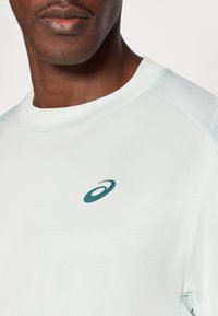 Αθλητική μπλούζα ανοιχτοπράσινου χρώματος με στρογγυλεμένο λαιμό, υφασμάτινες λεπτομέρειες δίχτυ στην περιοχή των ώμων και μικρό σκούρο πράσινο λογότυπο στο στήθος.