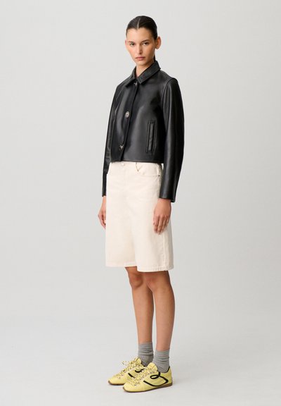 Mujer con chaqueta de cuero negra, falda beige hasta la rodilla, calcetas grises y zapatillas amarillas, de pie frente a un fondo claro y liso.