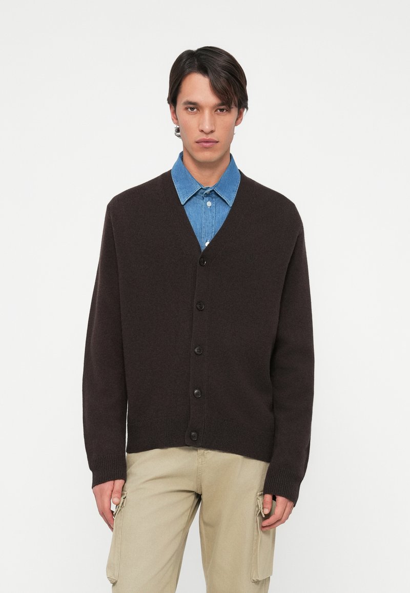 Brun knäpp-up cardigan tillverkad av mjukt stickat material, med V-hals, ribbade ärmslut och enkel design bärs över en blå denimskjorta.