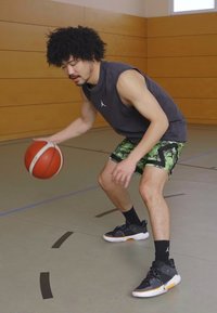 Μπάσκετ παίκτης με γκρι αμάνικο μπλουζάκι και πράσινα καρό σορτς, ντριμπλάροντας μια κόκκινη μπάλα μπάσκετ φορώντας μαύρα αθλητικά παπούτσια.