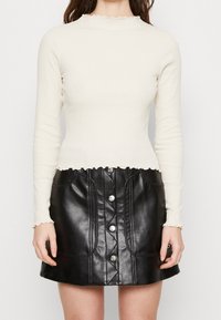 Krämfärgad långärmad topp med vågformad halsringning, parat med en hög midja svart läder minikjol med silverknappar och sömnadsdetaljer.