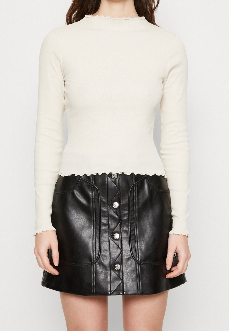 Krämfärgad långärmad topp med vågformad halsringning, parat med en hög midja svart läder minikjol med silverknappar och sömnadsdetaljer.