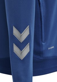 Blauwe stoffen mouw met twee reflecterende chevronstrepen en een ritssluitingzak op een getextureerd blauw kledingstuk met de tekst "BEECOOL".