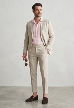 Mand stående indendørs iført en beige jakkesæt, lyserød skjorte, brune loafers, holder solbriller i den ene hånd, ser let til siden.