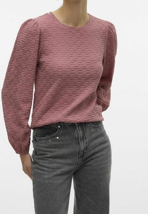 Frau trägt eine strukturiert roséfarbene, langärmelige Bluse mit aufgepufften Schultern, kombiniert mit hoch taillierten schwarzen Jeans mit kleinen Verzierungen.