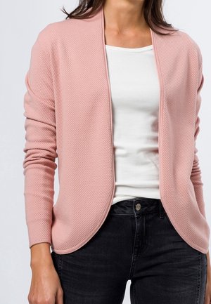 Femme portant un cardigan rose texturé ouvert sur le devant, un t-shirt blanc ajusté et un jean noir, montrée des épaules aux hanches.