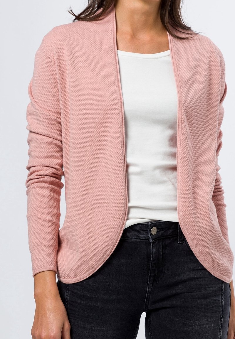 Femme portant un cardigan rose texturé ouvert sur le devant, un t-shirt blanc ajusté et un jean noir, montrée des épaules aux hanches.