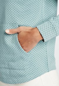 Világoskék, hosszú ujjú felső fekete pöttyökkel, elülső zsebbel és hajtott ujjakal, texturált, rugalmas anyagból készült.