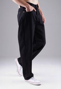 Schwarze Jeanshose mit lockerer Passform, ausgestattet mit Fronttaschen, kupferfarbenen Hardware-Akzenten und kombiniert mit weißen Turnschuhen.