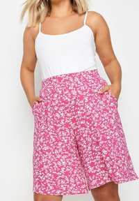 Woman wearing white sleeveless top and high-waisted pink floral shorts with pockets, standing with hands in pockets.