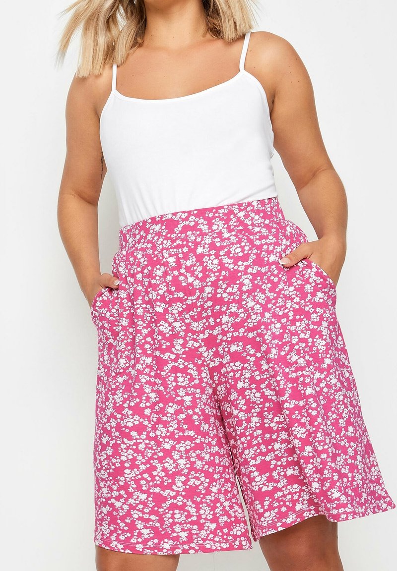 Woman wearing white sleeveless top and high-waisted pink floral shorts with pockets, standing with hands in pockets.