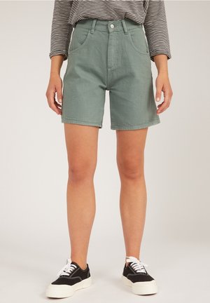 Pantaloni corti in denim verde di cotone con cinque tasche, chiusura con bottone e orlo leggermente arrotolato, abbinati a sneaker nere su uno sfondo chiaro.