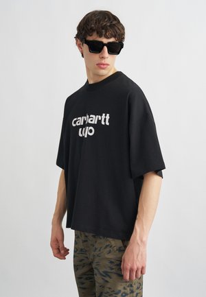 Młody mężczyzna w czarnej, oversize'owej koszulce Carhartt, czarnych okularach przeciwsłonecznych i zielonych spodniach w wzory, stoi na tle jednolitego jasnego tła.