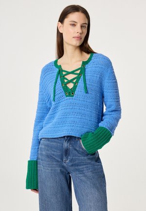 Frau trägt einen blauen Strickpullover mit grün geschnürtem Ausschnitt und Manschetten, kombiniert mit hoch taillierten blauen Jeans, steht mit einer Hand in der Tasche.