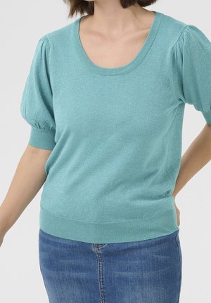 Vrouw draagt een lichtblauwe, korte mouw trui met pofmouwen en een denim rok tegen een effen lichte achtergrond.