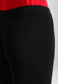 Zwarte op maat gemaakte broek met een gladde textuur, voorzien van een eenvoudige achterzak en een traditionele tailleband, gecombineerd met een rode top.