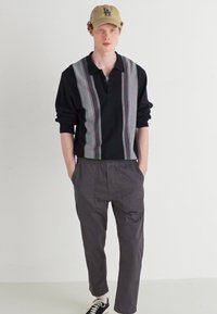 Crna polo majica dugih rukava s vertikalnim prugama, uparena sa sivim tapered hlačama i crnim tenisicama. Bež kapa upotpunjuje outfit.