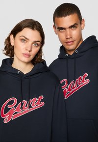Mørkeblå hettegensere med en rød "Gant"-logo, featuring snorer og en frontlomme. Glatt tekstur og uformelt unisex design.