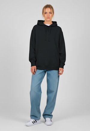 Felpa nera con cappuccio e tasca frontale, abbinata a jeans larghi azzurri e scarpe bianche con strisce nere. Design semplice, vestibilità casual.