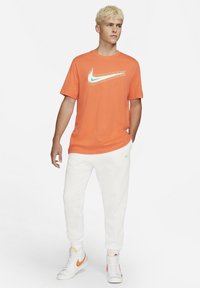 Orange cotton t-shirt featuring a blue and yellow Nike logo, paired with white sweatpants and white high-top sneakers with orange accents.