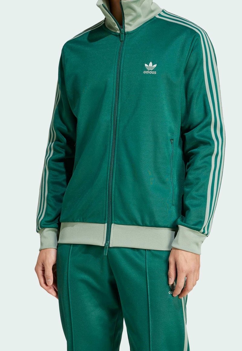 Grön sportoverall med zippered jacka med vita ränder och logotyp, kombinerad med matchande byxor. Slät tyg med ribbade muddar.